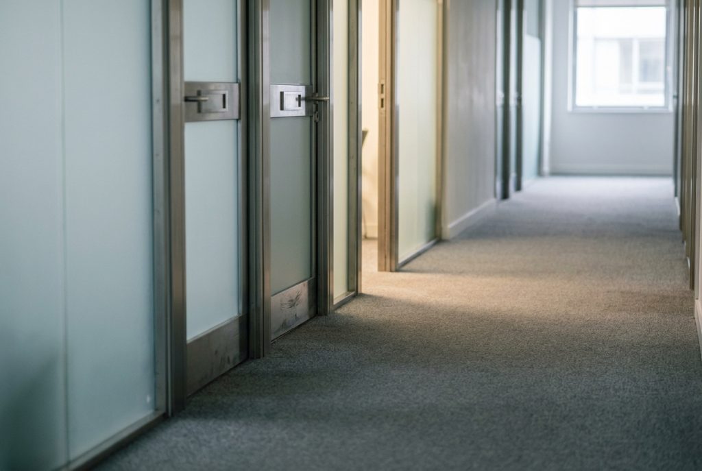 Editorial photograph of a quiet British office corridor with frosted glass office doors, a door partway down the corridor partly open with warm light spilling out, suggesting the named, accountable role of a Virtual Compliance Director.